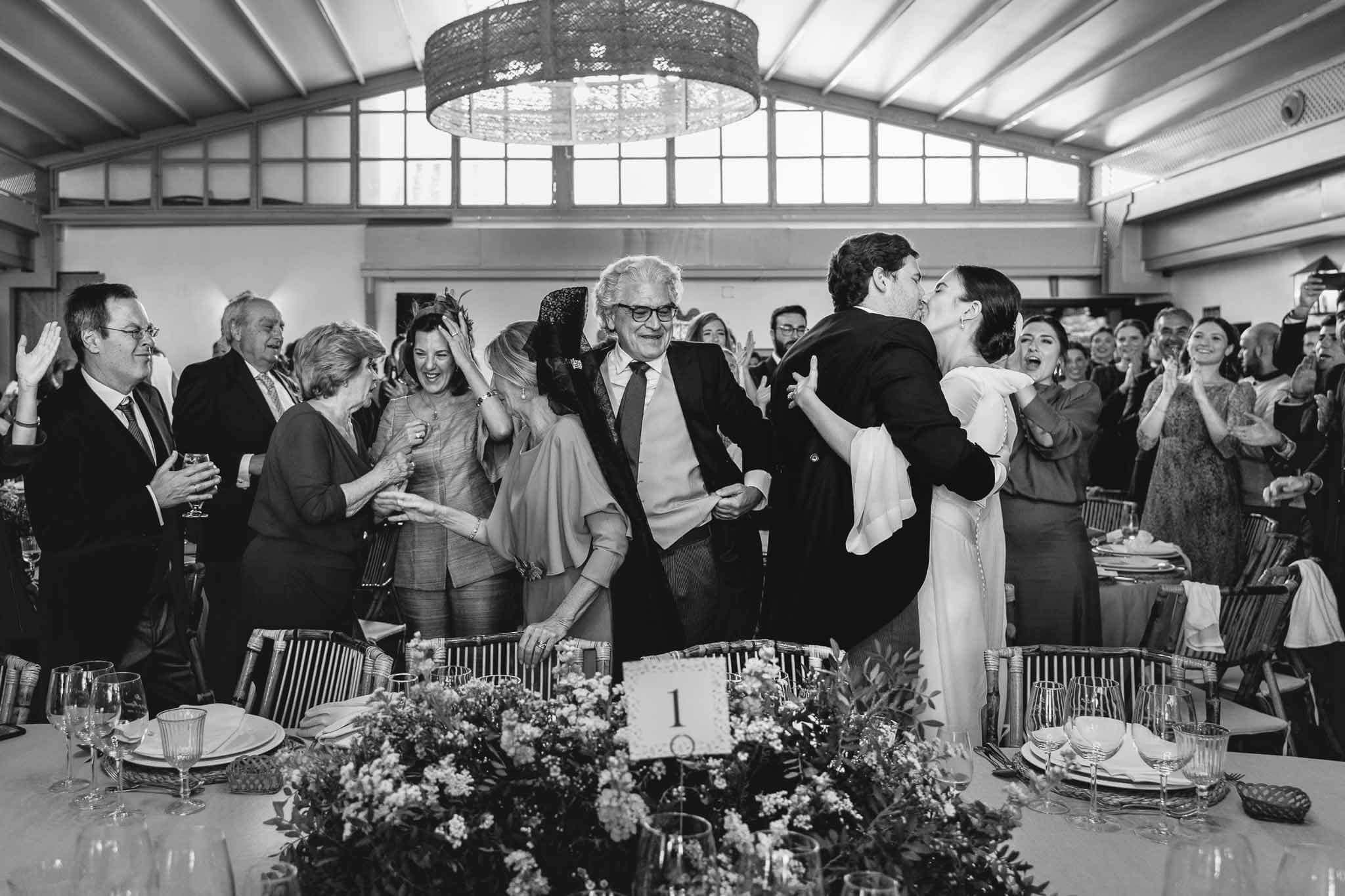 Novios besándose en la mesa presidencial de su boda en compañía de personas invitadas (Foto en color)